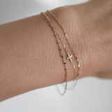 Model wearing three layered 14K gold bracelets on wrist, including a cross charm, fine bead chain, and rectangular link design