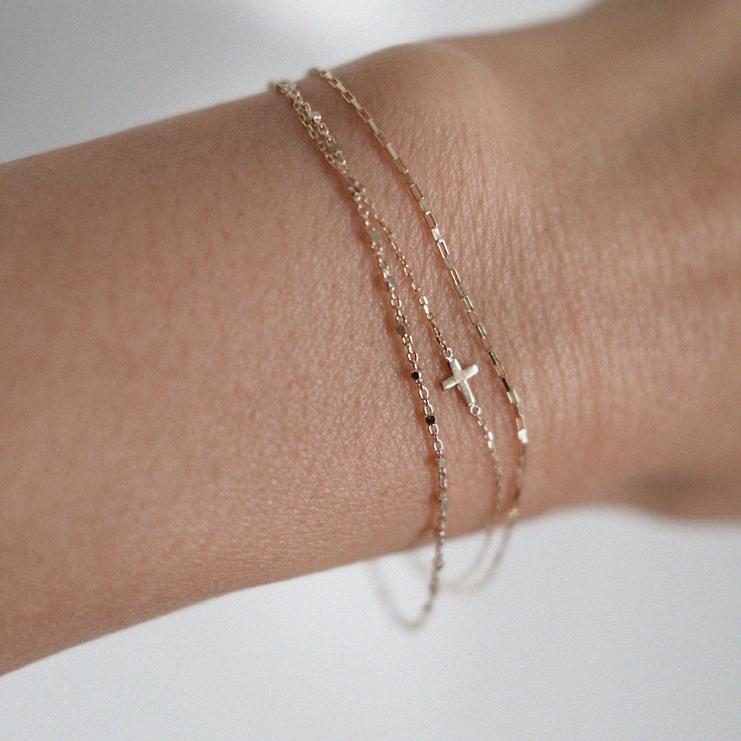 Model wearing three layered 14K gold bracelets on wrist, including a cross charm, fine bead chain, and rectangular link design