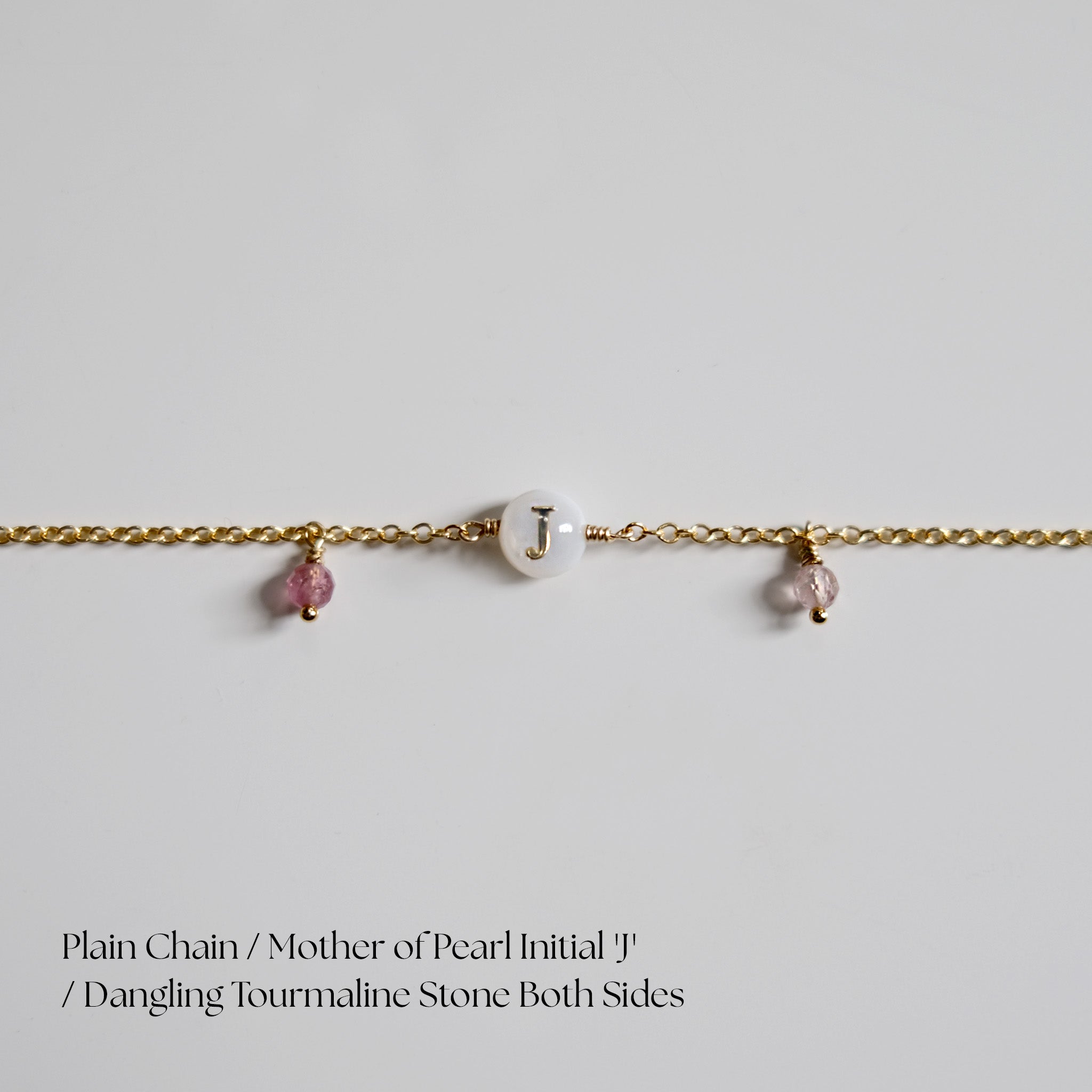 Gold chain bracletlet with mother of pearl initial 'J' and dangling tourmaline stones on a light gray background.