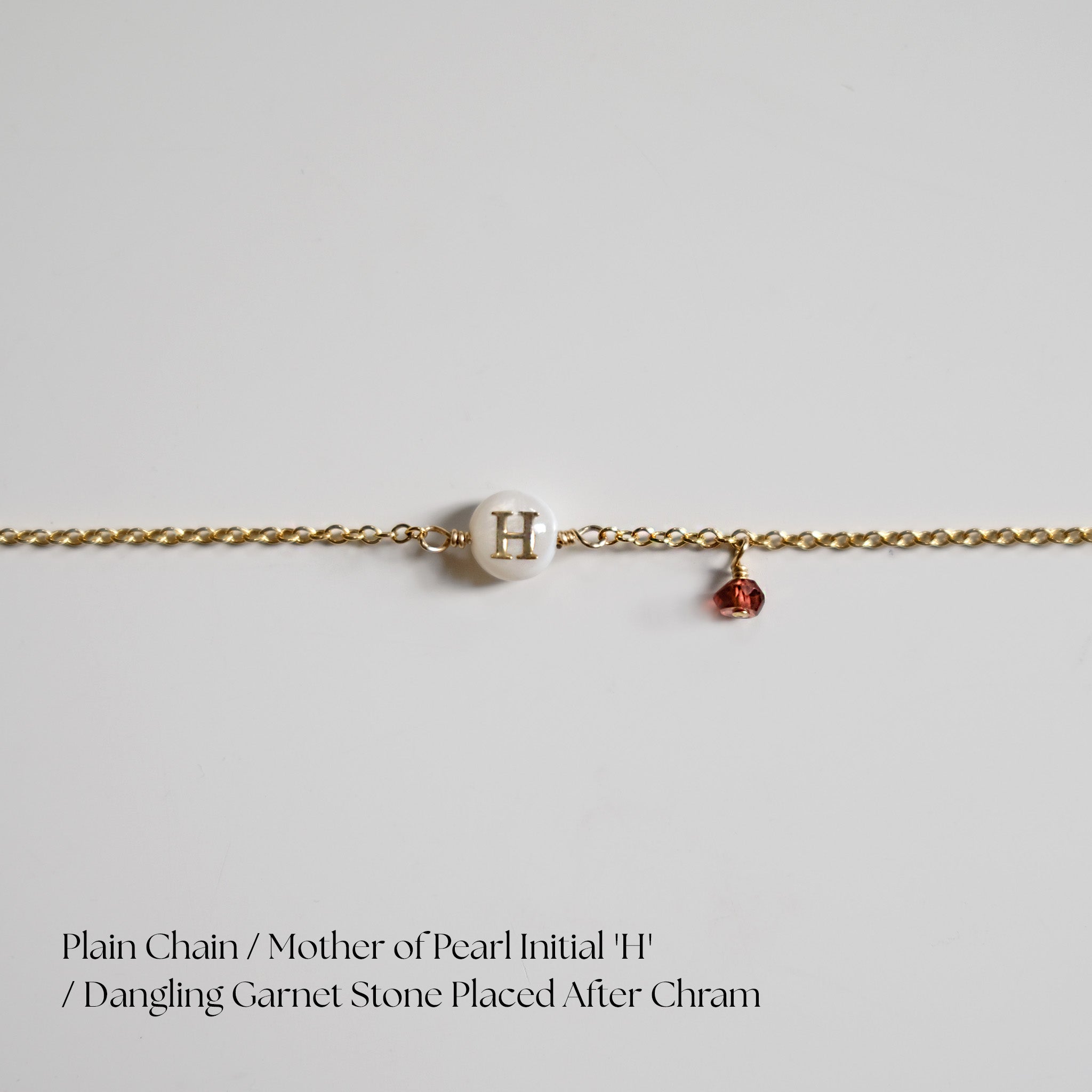 Gold chain bracelet with a mother of pearl initial 'H' and garnet stone on a light gray background