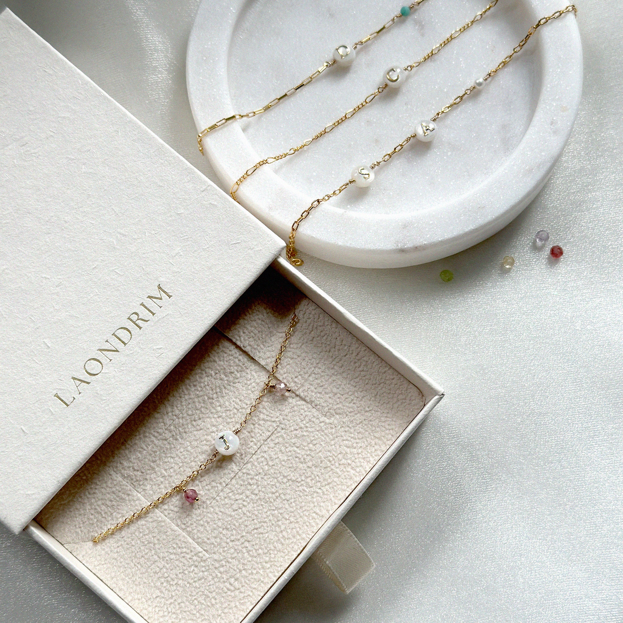 Gold bracelet with initial charms and gemstones on a white tray and gift box with 'LAONDRIM' branding.