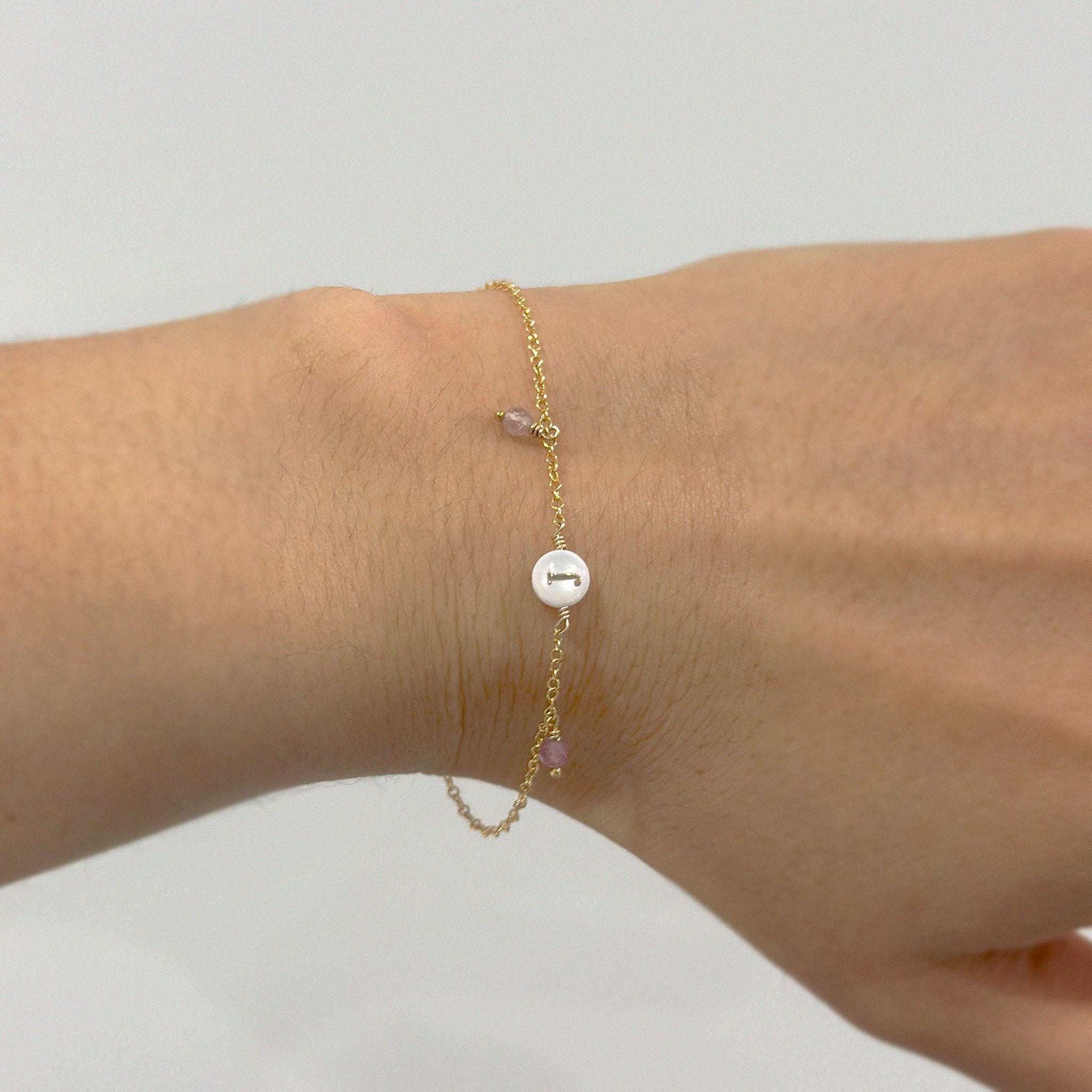 Gold bracelet with a mother of pearl initial charm and small tourmaline gemstone on a wrist against a neutral background