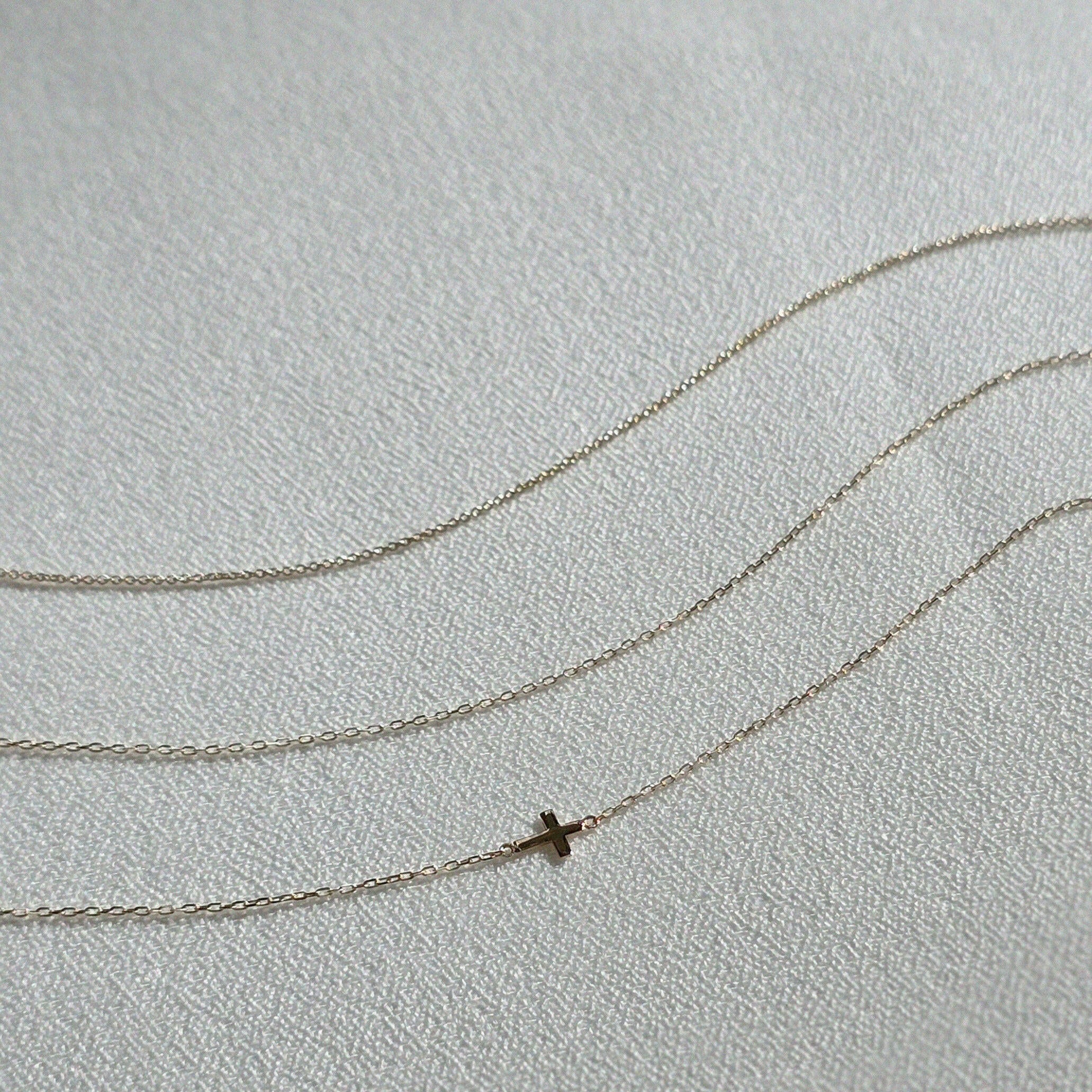 Three dainty 14K gold chain bracelets displayed on a soft textured fabric, including one with a small cross charm