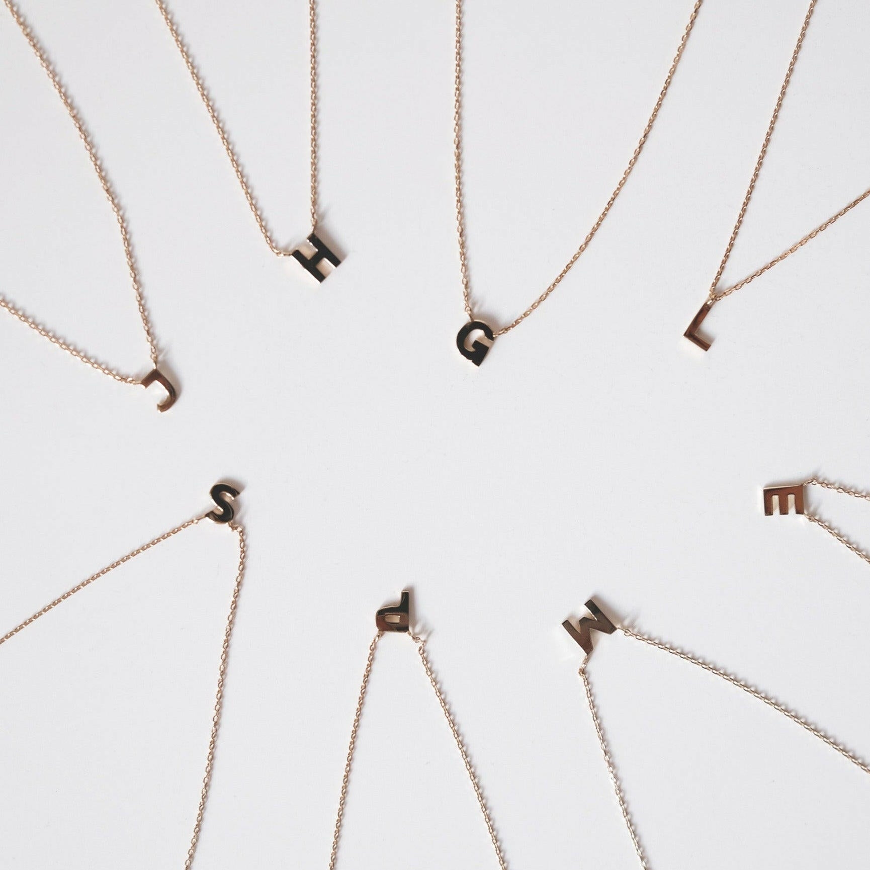 Multiple initial necklaces, including J, H, G, L. E, M, P and S, displayed with their chains against a clean white backdrop