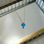 Blue opal cross-shaped pendant on a gold chain against a neutral background