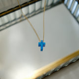 Blue opal cross-shaped pendant on a gold chain against a neutral background