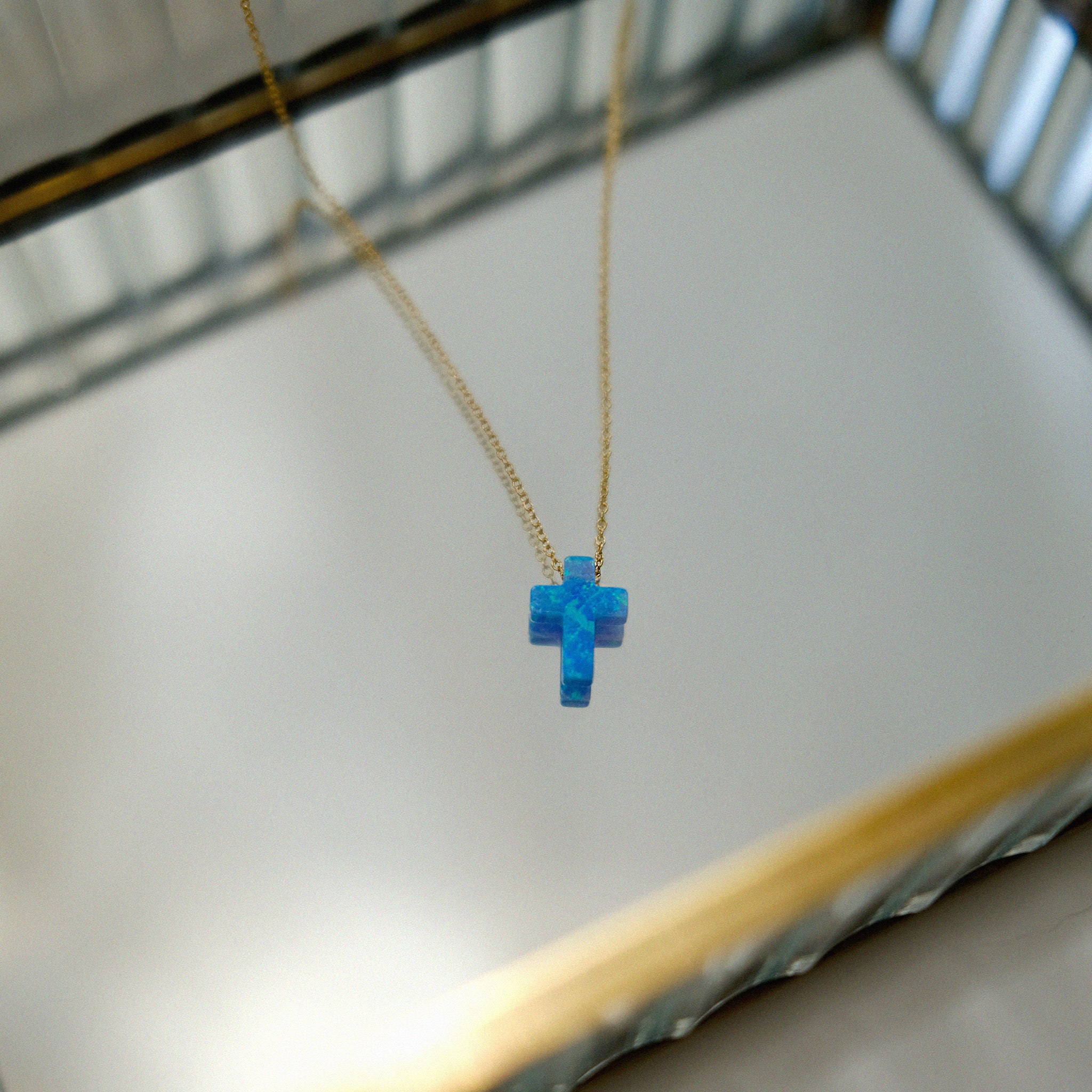 Blue opal cross-shaped pendant on a gold chain against a neutral background