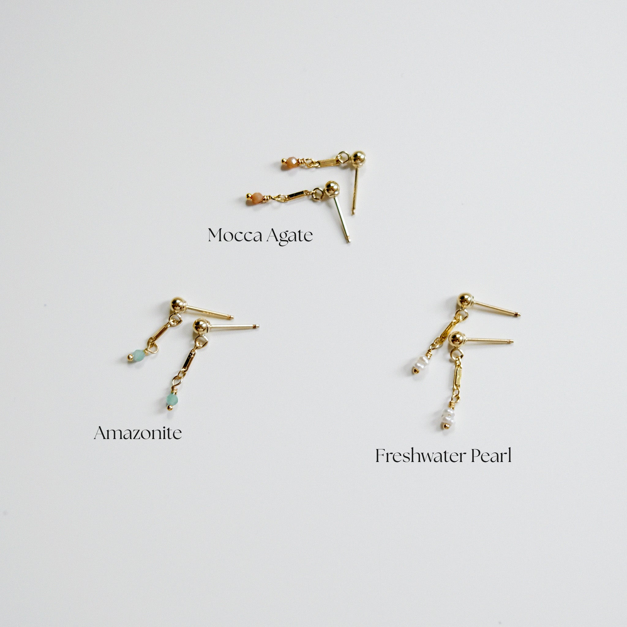 Three gold earrings with gemstones labeled 'Moecca Agate', 'Amazonite', and 'Freshwater Pearl' on a light gray background.