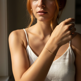 Woman wearing a white top with a gold chain with a small bar bracelet, sitting in soft light.