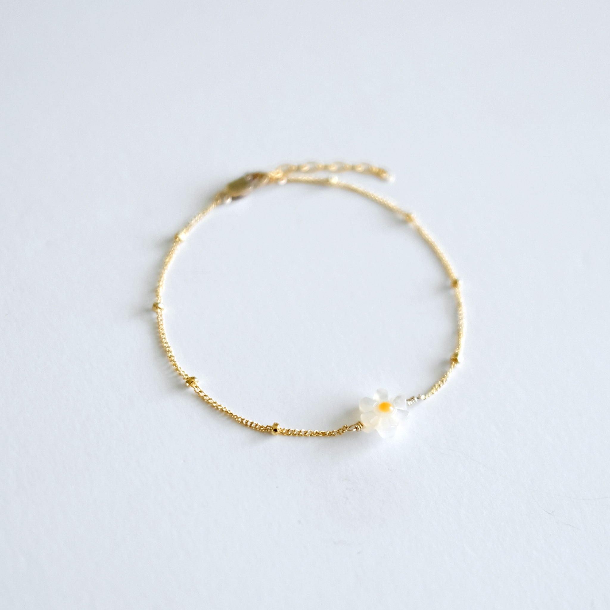Gold bracelet with a small mother of pearl daisy flower charm on a light gray background