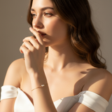 Woman in a white off-shoulder top with a neutral background, wearing a dainty gold chain with two pearl bracelet.