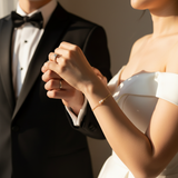 Close-up of a couple holding hands with wedding rings, dressed in formal attire. Women wearing a small bracelet with 2 pearls on dainty gold chain.