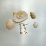 Gold earrings with starfish charms and pearls on a light background with shells.