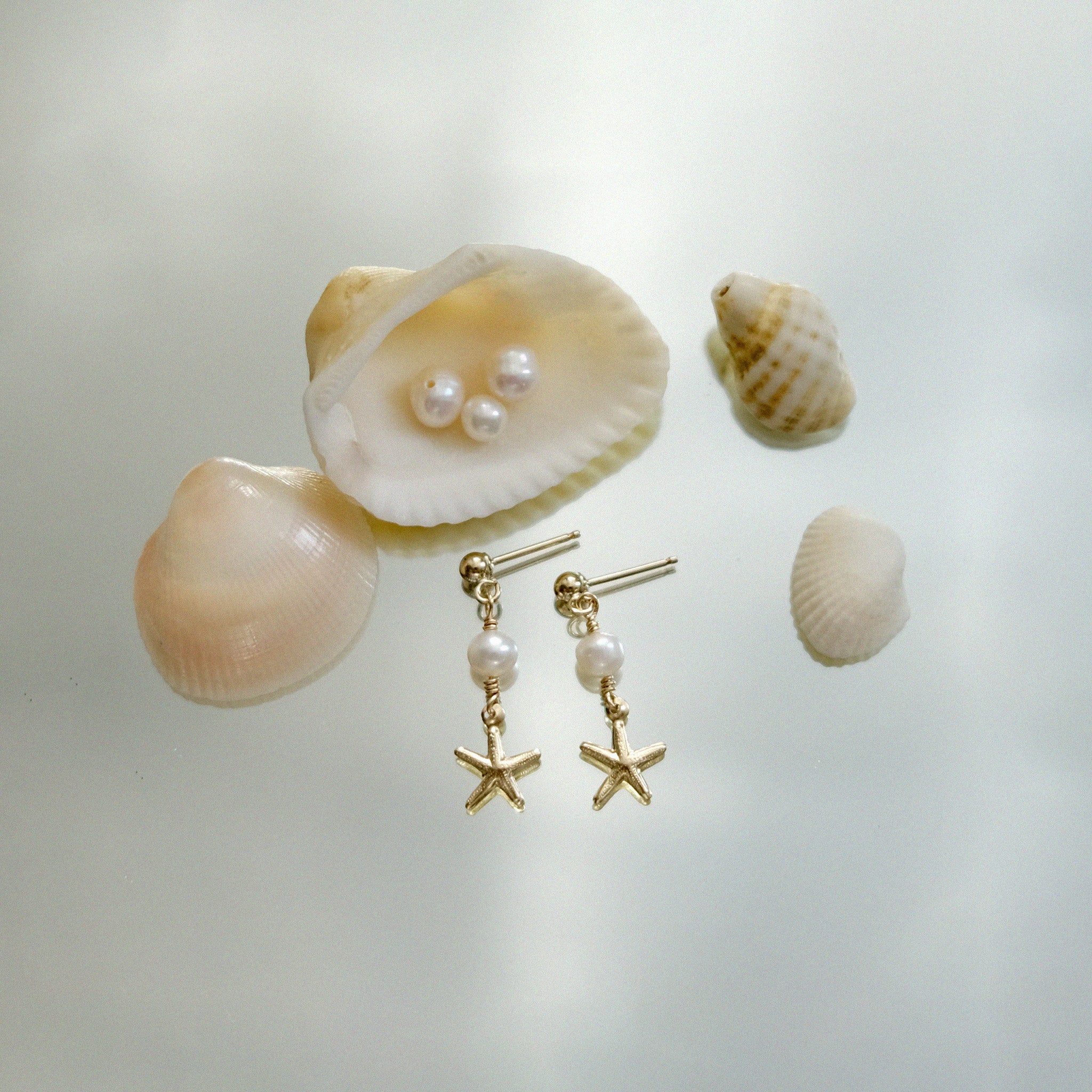 Gold earrings with starfish charms and pearls on a light background with shells.