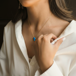 Person wearing a gold necklace with a blue pendant, touching their chest.