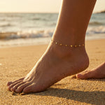 Gold anklet on a foot on a sandy beach with ocean waves in the background