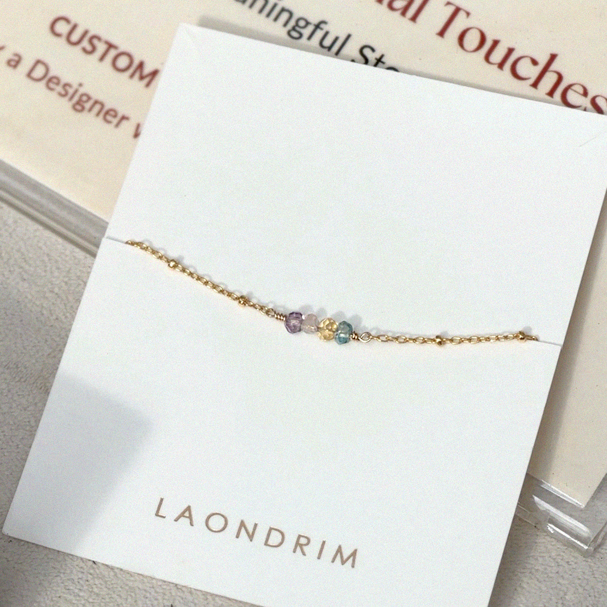Gold bracelet with multicolored beads on a white card with 'LAONDRIM' branding.