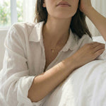 Woman wearing gold jewelry including a necklace and bracelet, sitting on a white couch.