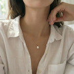 Person wearing a delicate gold necklace with a pearl pendant, set against a blurred indoor background.