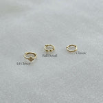 Side-by-side comparison of three 14K gold huggie hoops – Lil Clover with crystal flower, Ball Detail with gold spheres, and Classic plain hoop on soft fabric background