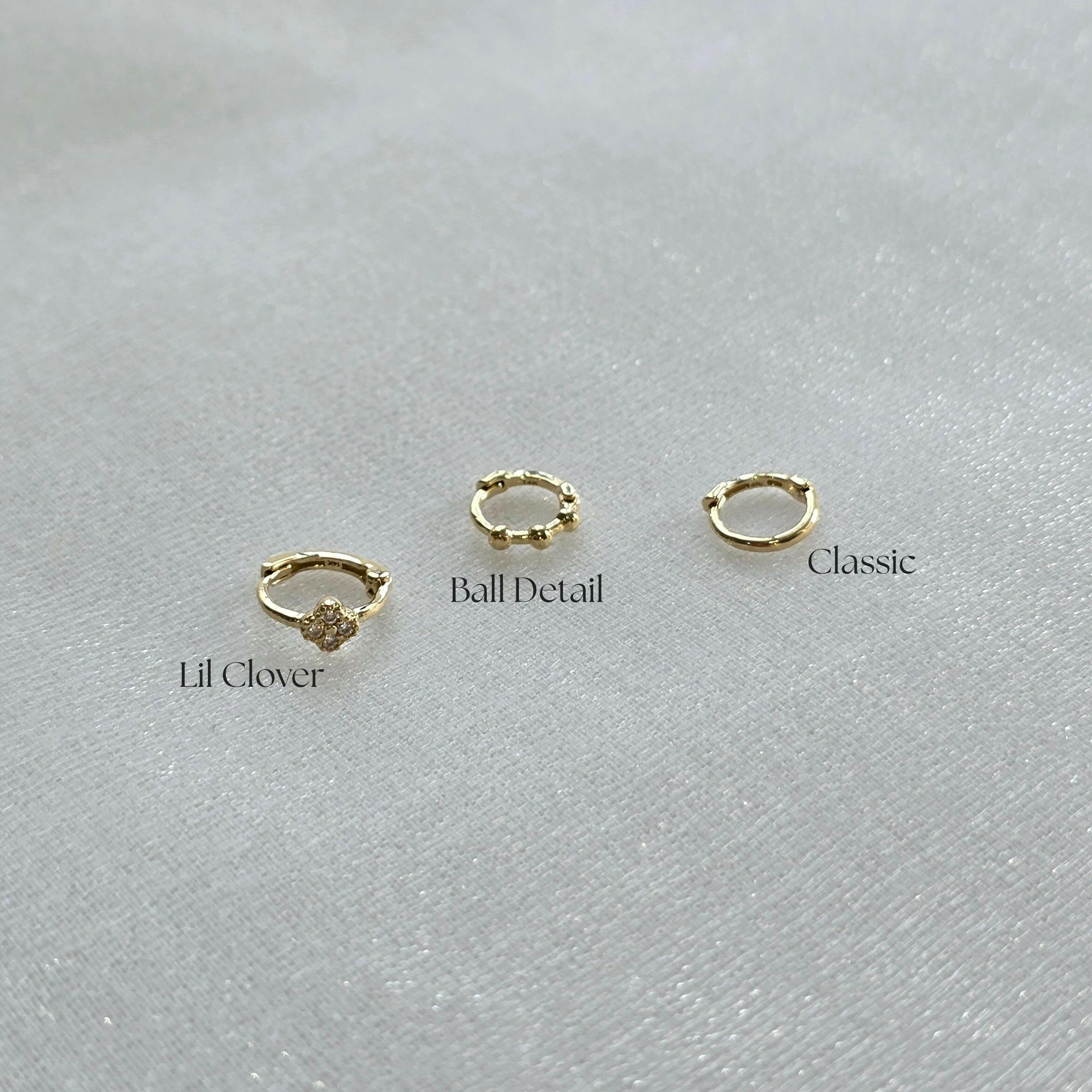 Side-by-side comparison of three 14K gold huggie hoops – Lil Clover with crystal flower, Ball Detail with gold spheres, and Classic plain hoop on soft fabric background