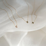 Three gold necklaces with letter pendants on a soft white fabric background