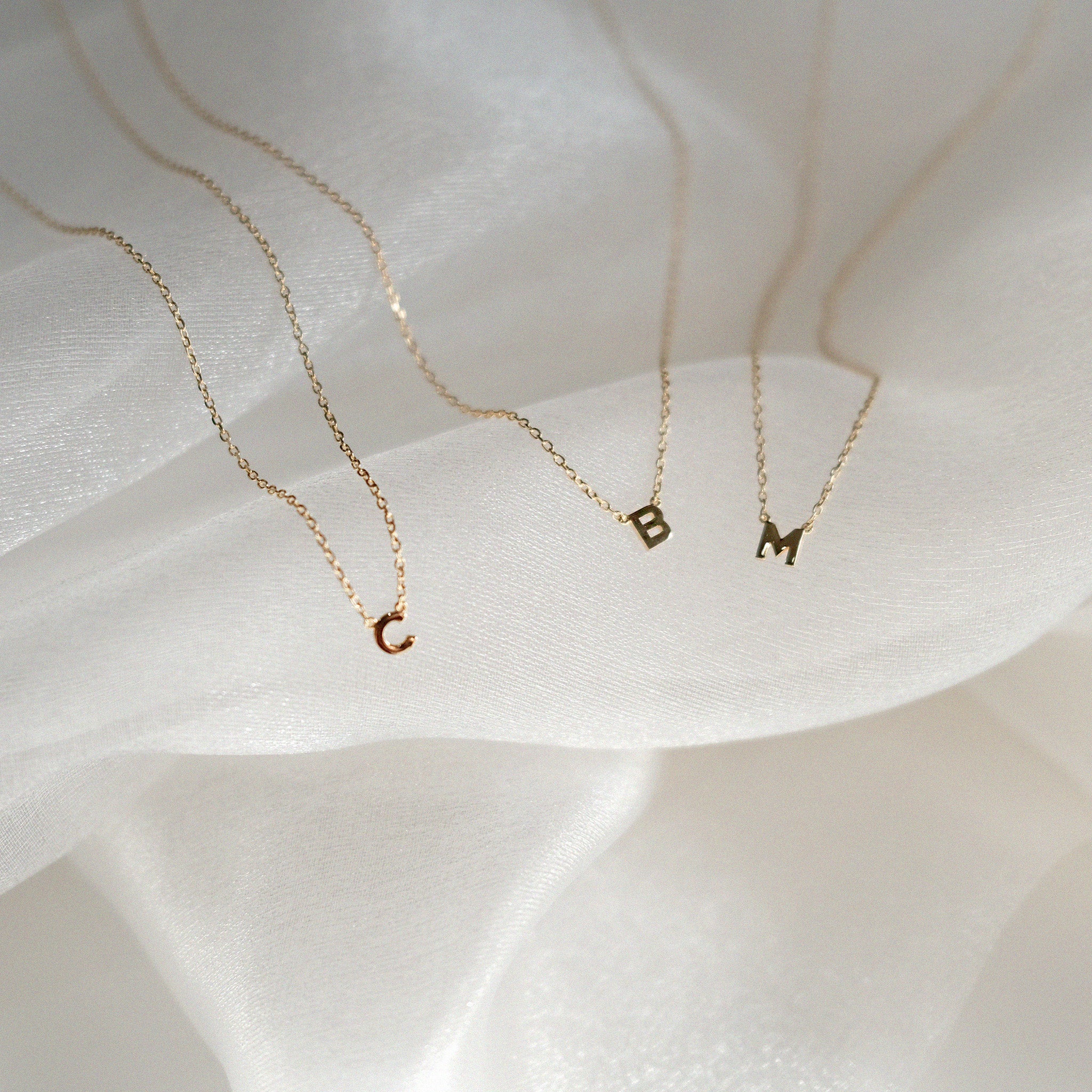 Three gold necklaces with letter pendants on a soft white fabric background