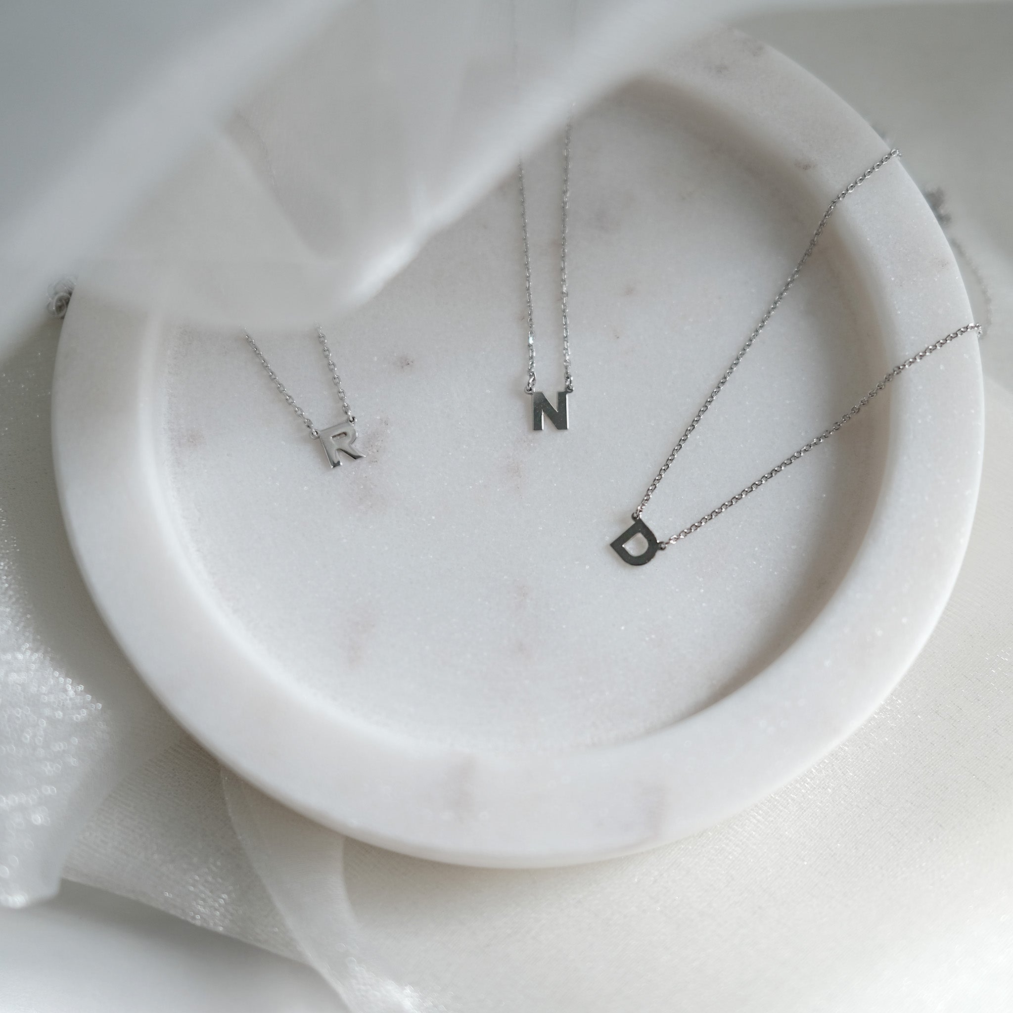 Three silver necklaces with initial pendants on a marble surface