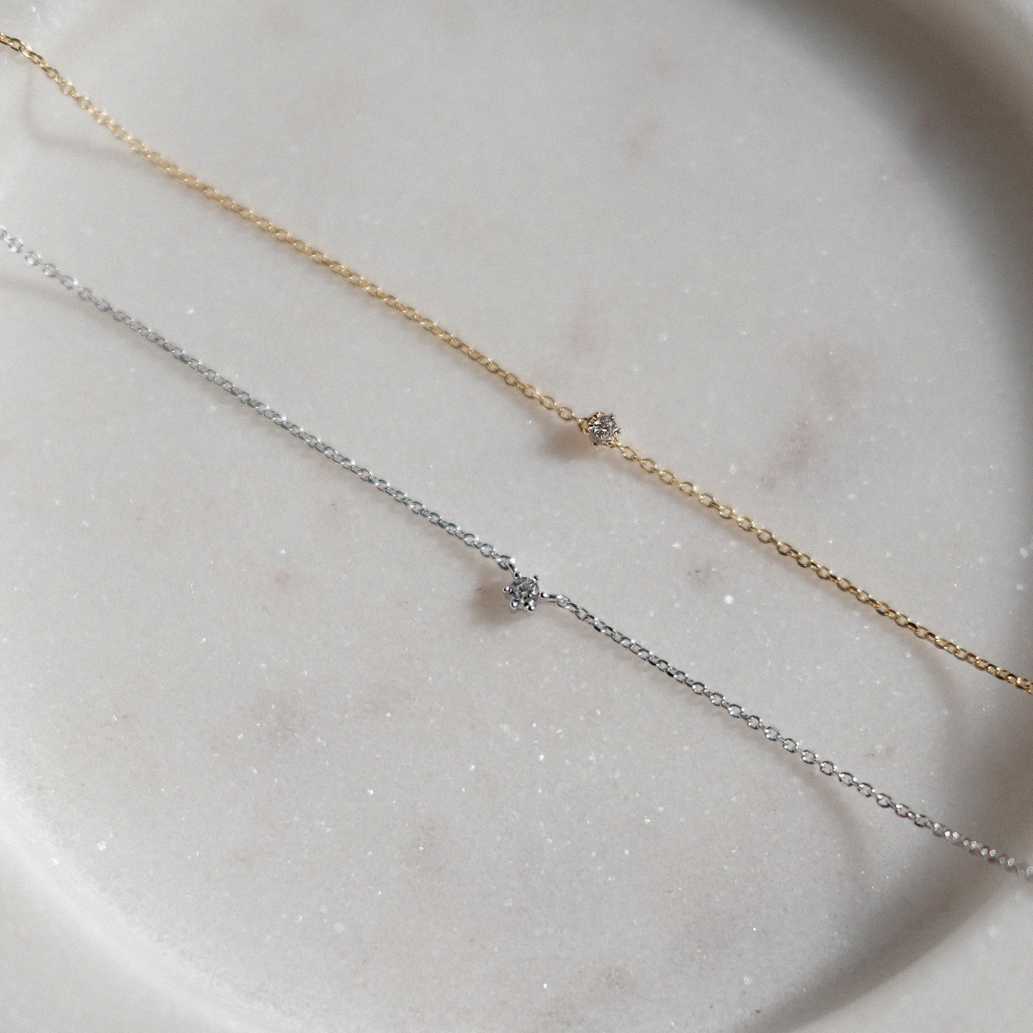 Two delicate necklaces, one gold and one silver, on a light stone surface.
